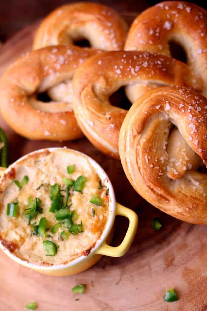 Overhead shot of pretzels next to a dish of beer cheese on wood cutting board with diced jalapeños. 