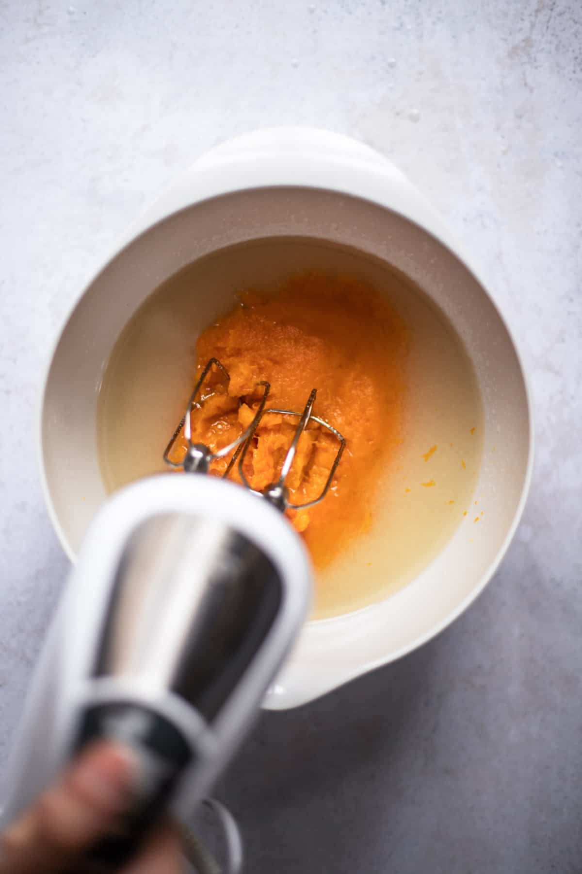 electric mixer beating the oil, sugar, and pumpkin together in white bowl.
