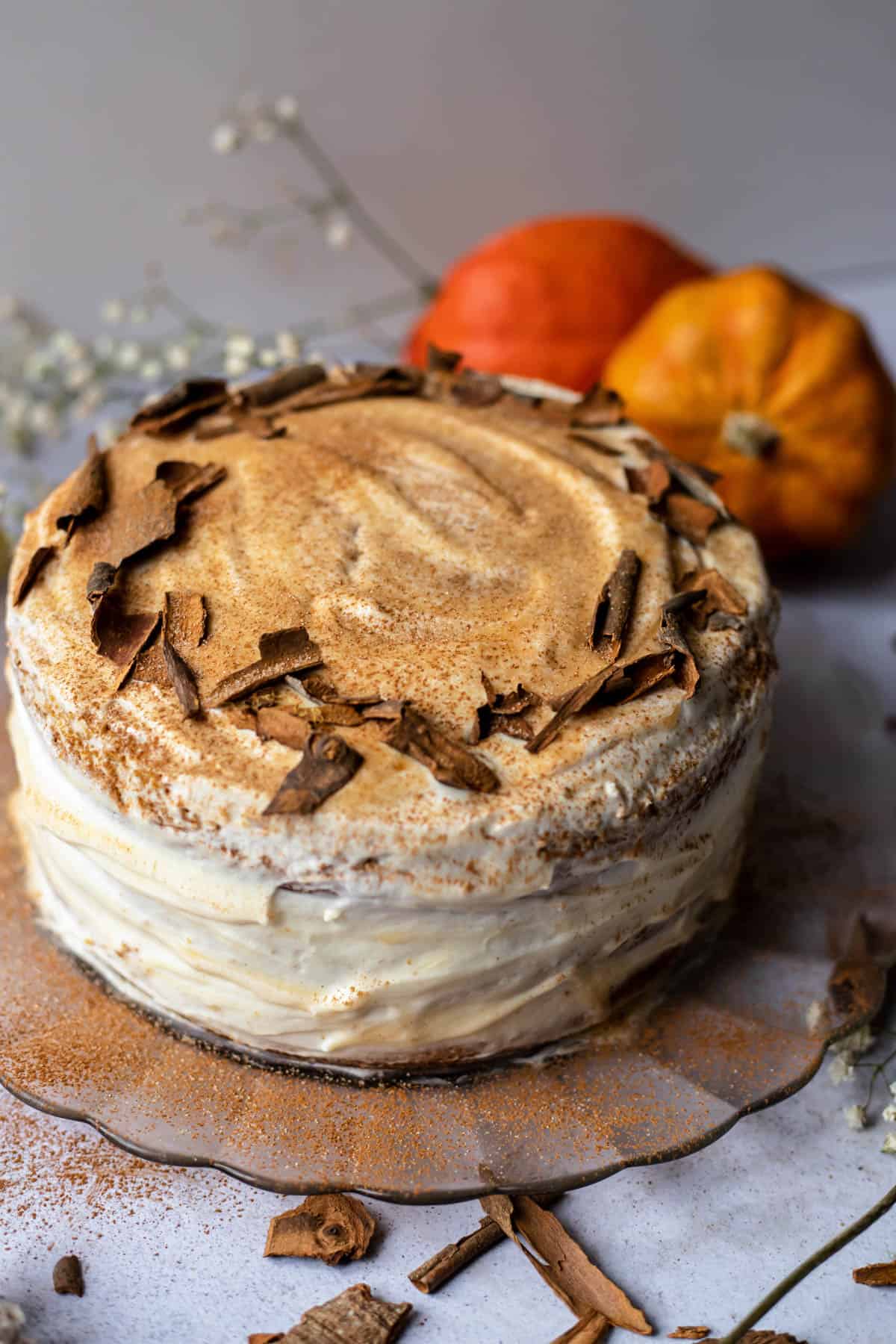 Whole frosted pumpkin cake on a wood cake plate with orange pumpkins in back. 