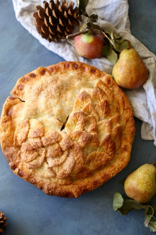 Whole Apple Pear Pie with double crust and a leaf crust garnish on a blue background with apples and pears around the pie. 