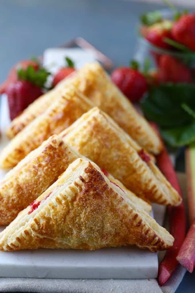 Stacked golden brown turnovers with stalks of fresh rhubarb and fresh strawberries around them. 