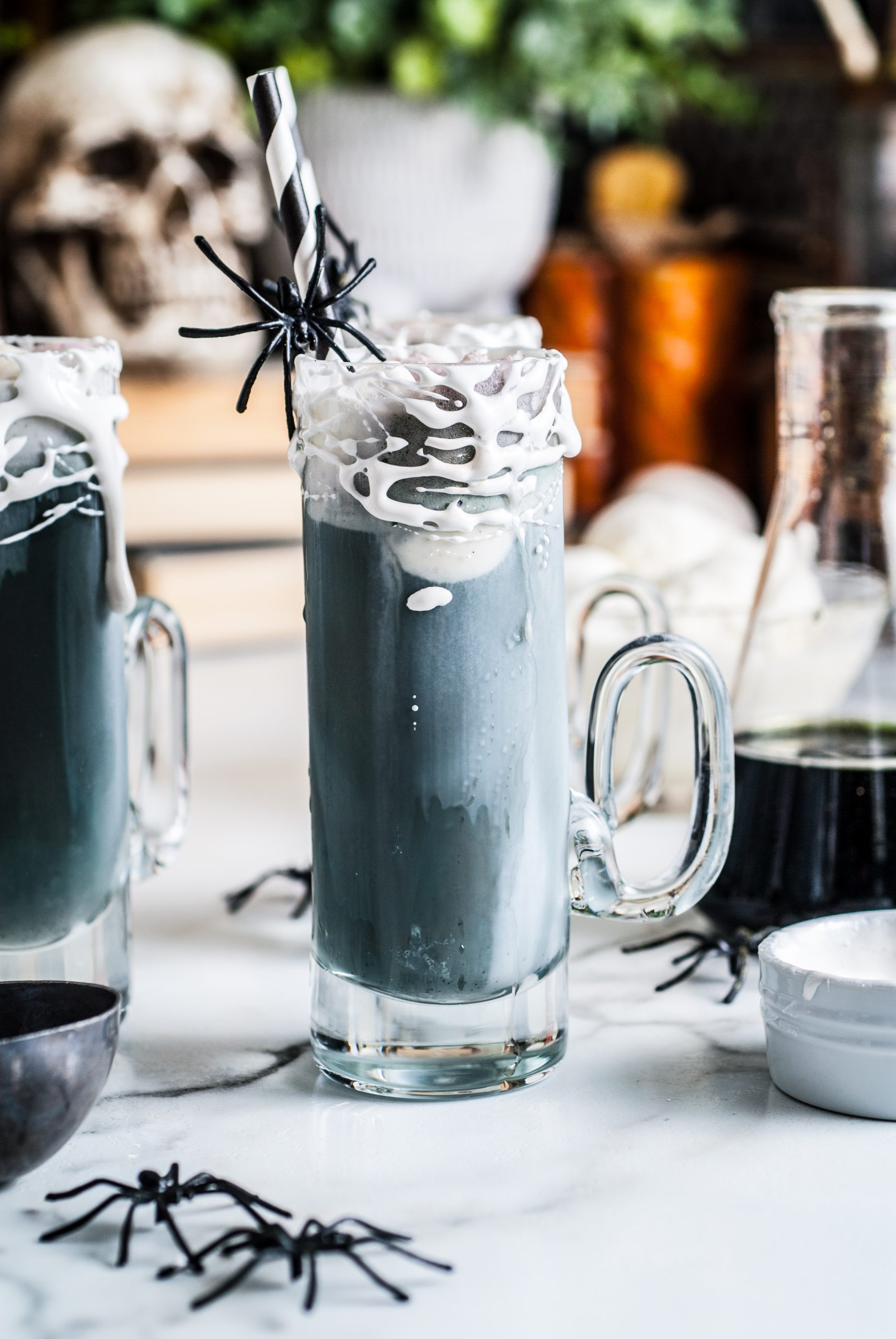 Shot of root beer float with skull in background and plastic spiders around glasses. 