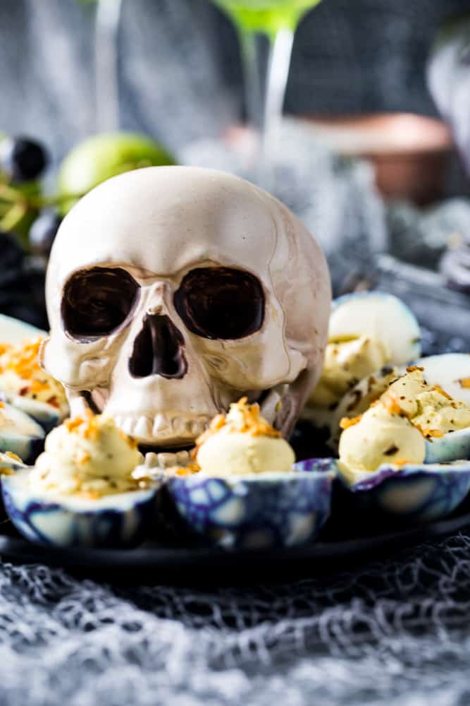 Spooky Deviled Eggs on a tray with a large skull in the center and black grapes in background. 