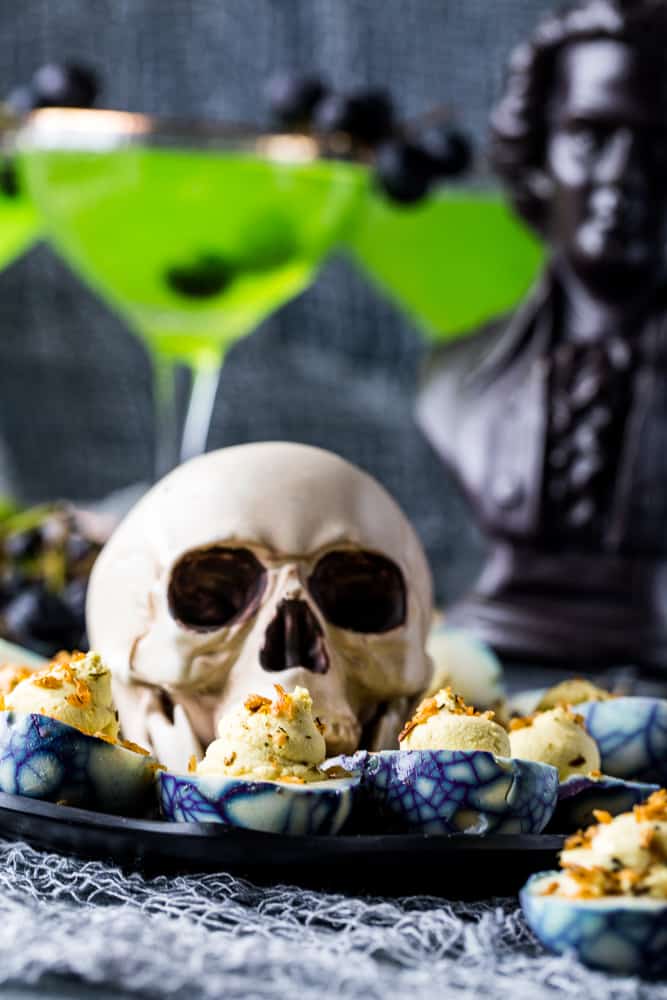Spooky Deviled Eggs on black platter with large plastic skull in center and green cocktails in the background. 