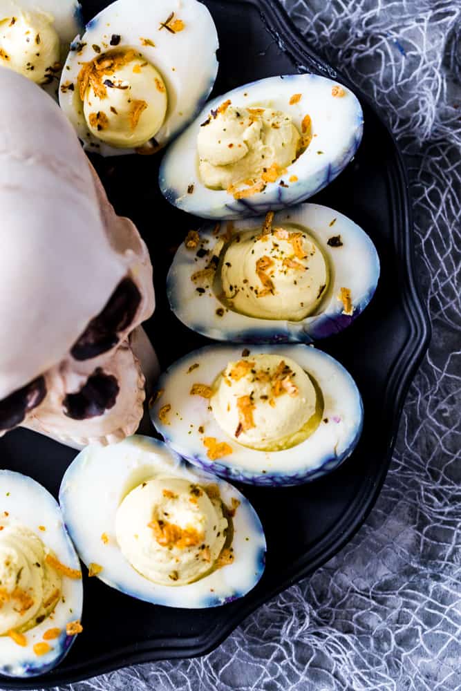 Overhead shot of the deviled eggs with crispy onions sprinkled on top on a black patter with skull in center. 