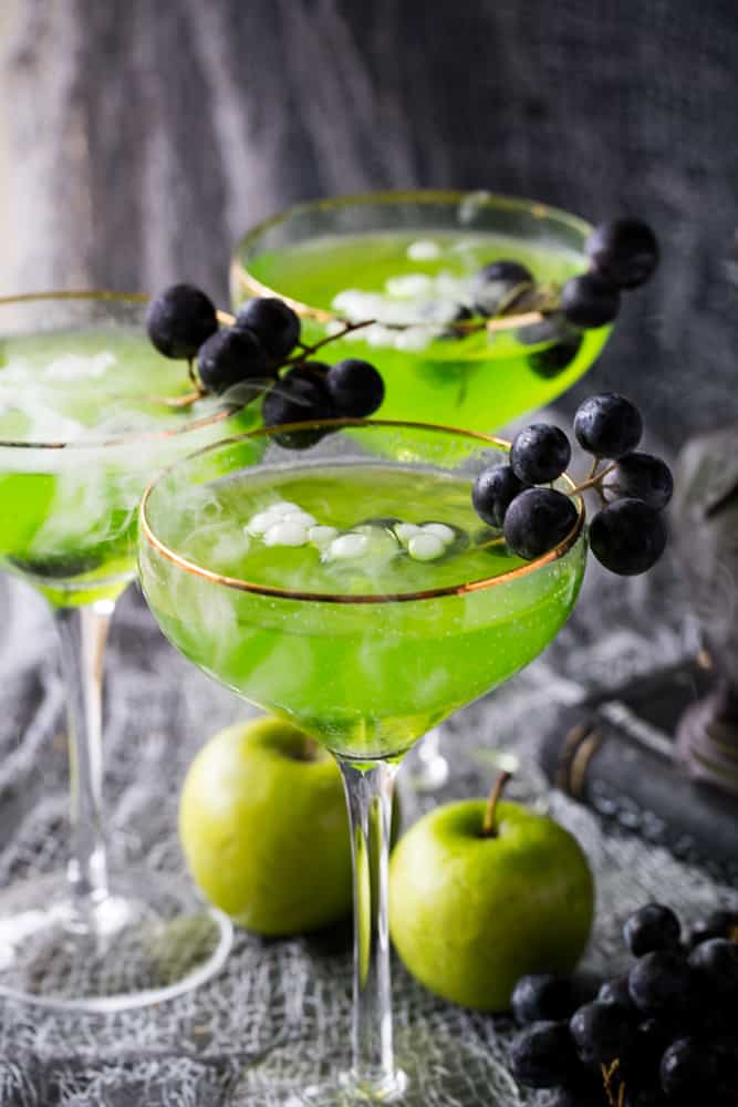 Glasses with green cocktail, dry ice, and black grapes on a grey background with green apples. 
