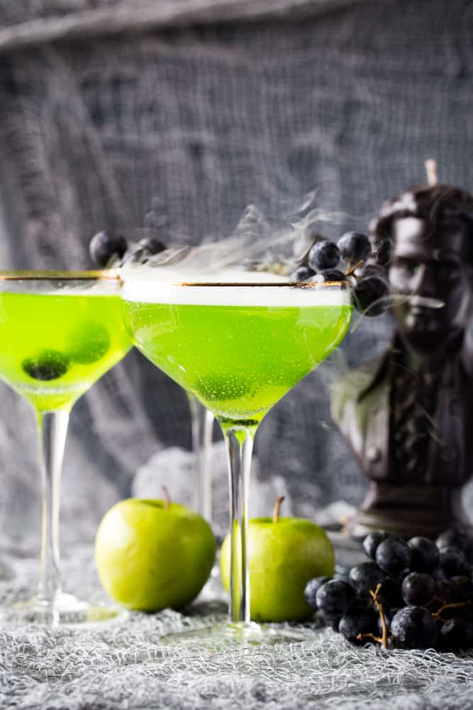 Poison Apple Cocktails on a grey background with black statue candle in the background. 