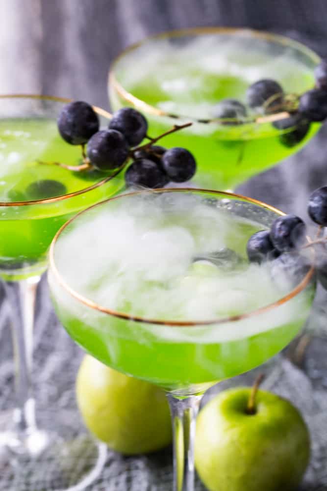 Three Poison Apple Cocktails that are garnished with black grapes and smoking because of the dry ice. 