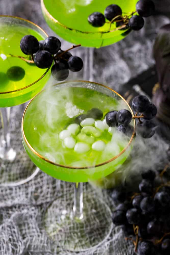 Overhead shot of 3 Poison Apple Cocktails with black grapes hanging off of the sides of the glasses. 