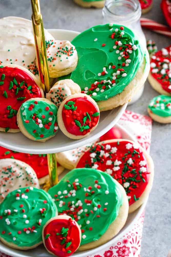 Small and large round cookies with red, green, and white frosting and sprinkles on a white tiered cookie plate. 