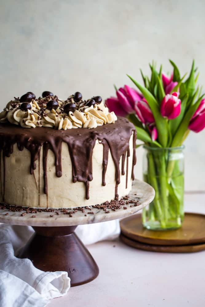 Fully assembled and decorated coffee layer cake with ganache and espresso beans on a brown marble cake plate with fresh pink tulips in the background. 