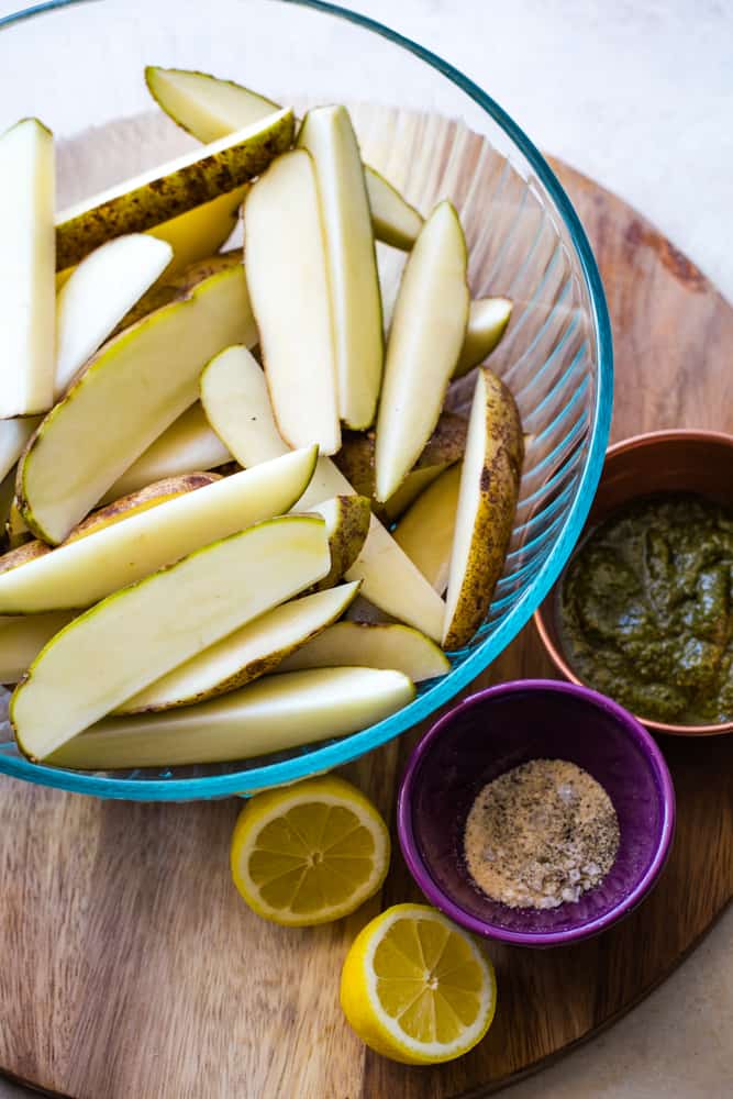 Large glass bowl of potato wedges with small bowl of pesto and spices on cutting board. 