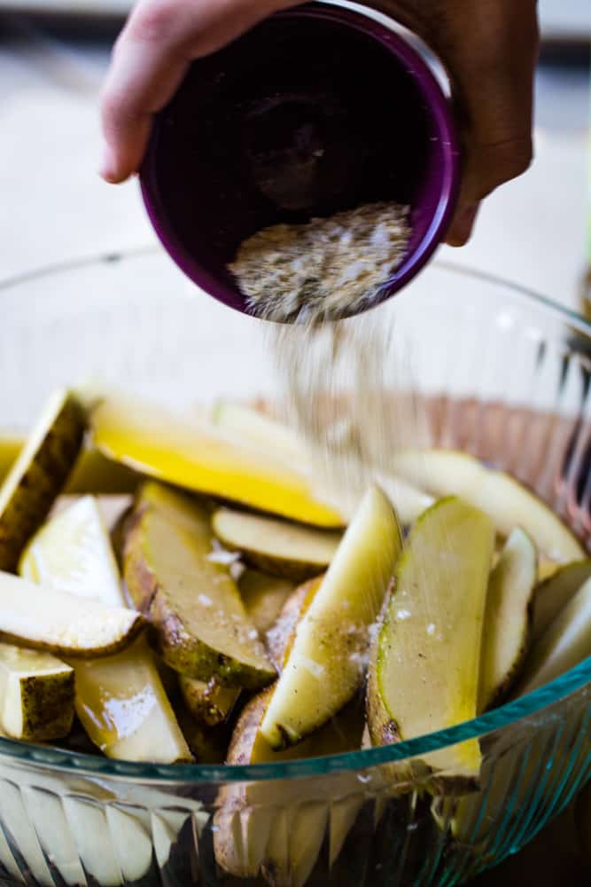 Hand pouring bowl of spices into the large bowl of potato wedges. 