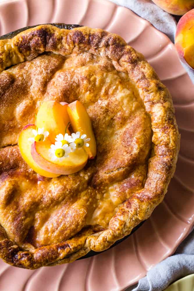 Double crust caramel peach pie on peach pie plate with whole peaches in the background. 