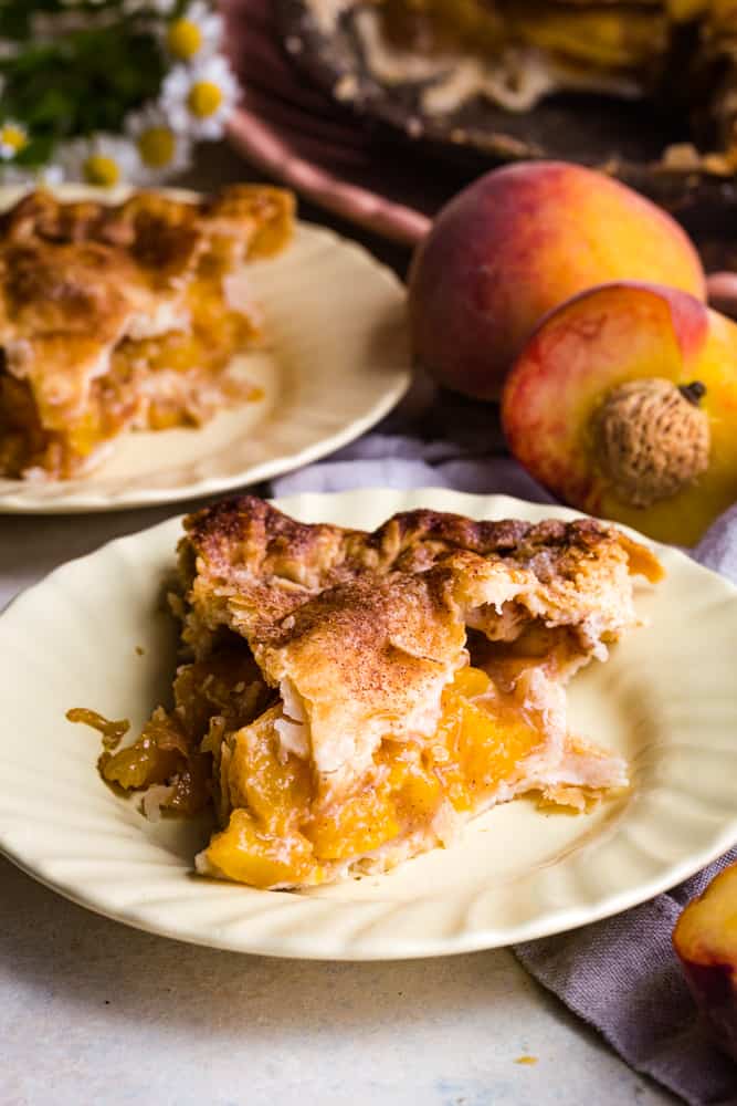 Slice of pie on yellow plate with a half a peach and pie in the background. 