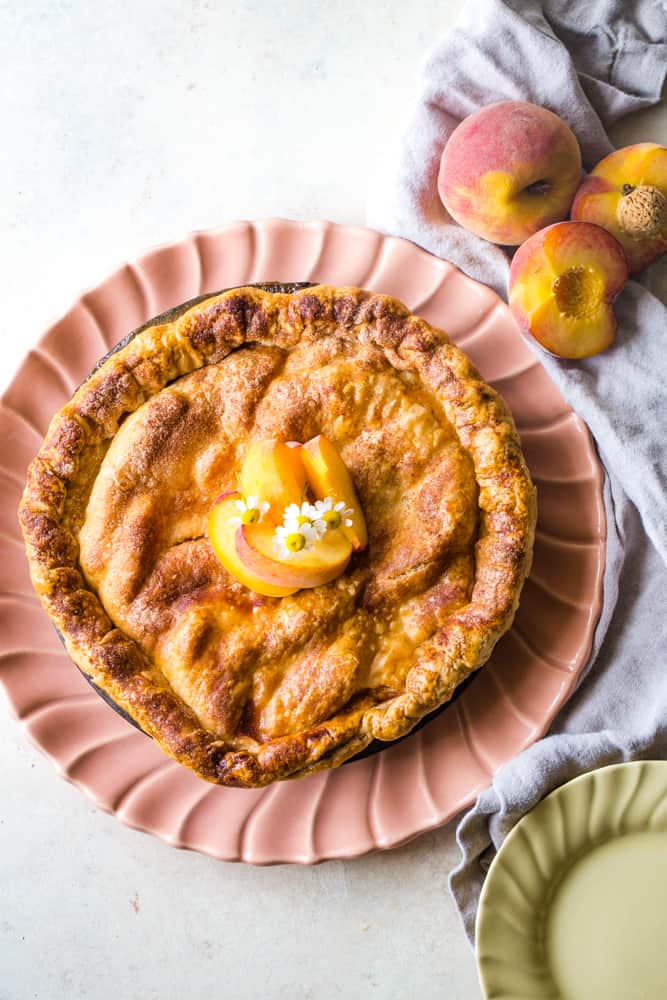 Overhead shot of entire pie on peach plate with decorative edges. Flowers and peach slices on top of pie. 