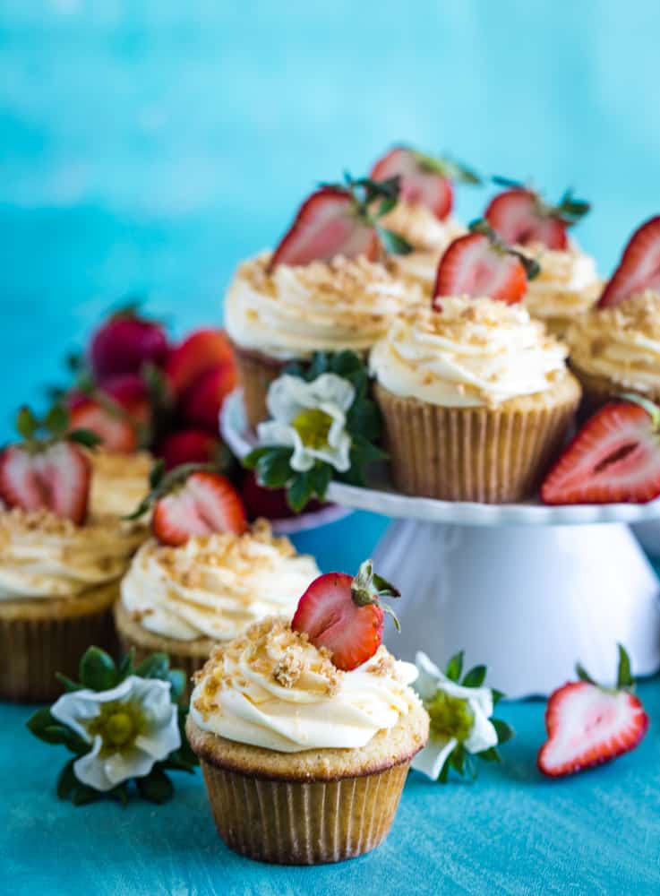 Cupcakes on white cake plate with more cupcakes in the foreground and strawberry blossoms. 