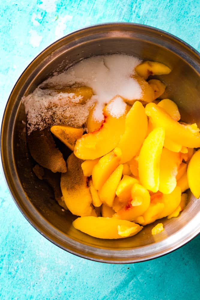 Sliced peaches, sugar, and cinnamon in a large metal bowl on blue background. 