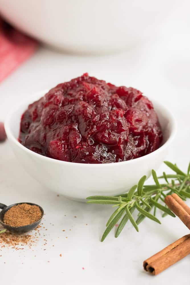 Small white bowl of leftover cranberry sauce for coffee cake with cinnamon sticks and rosemary next to it. 