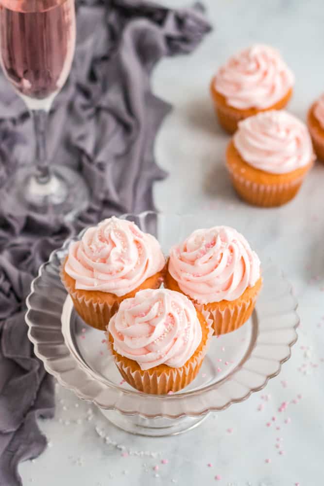 3 pink champagne cupcakes on clear cake stand with purple dish towel