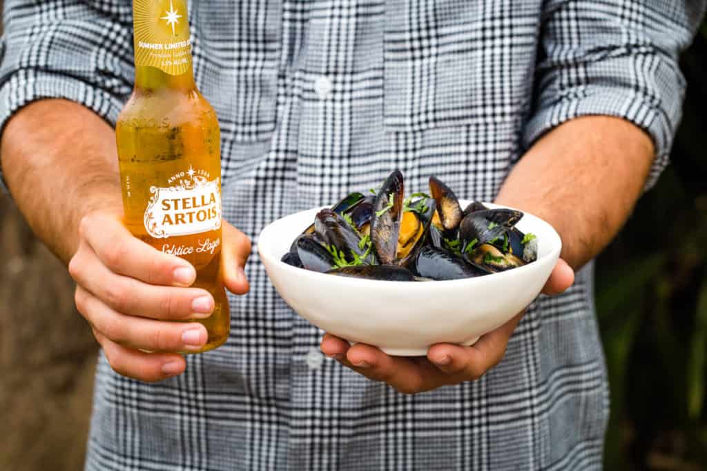 hands holding a bottle of stella artois and a white bowl of mussels