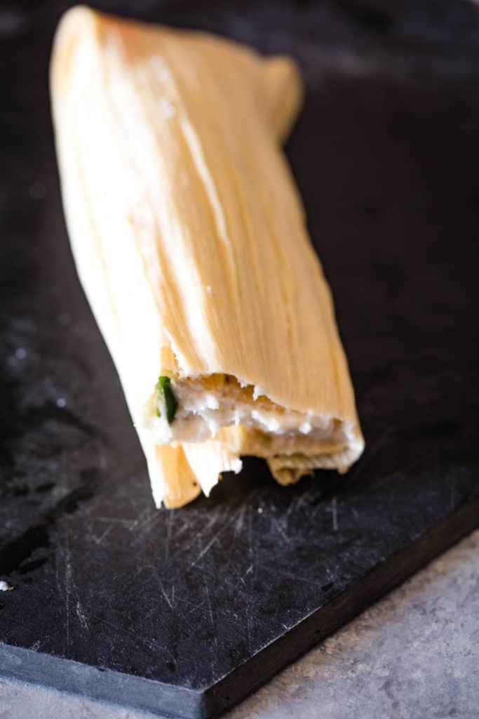 rolled corn husk with uncooked tamale on black cutting board
