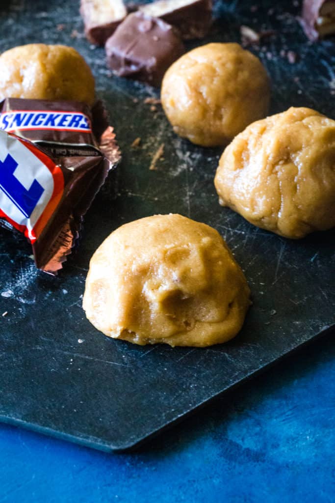 Snickers stuffed dough balls on black cutting board with candies around it. 