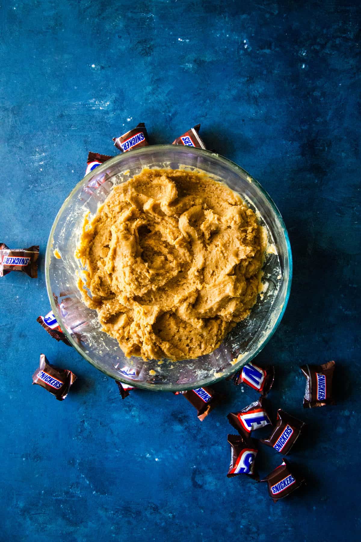 Bowl of peanut butter cookie dough on blue background with snickers around it. 