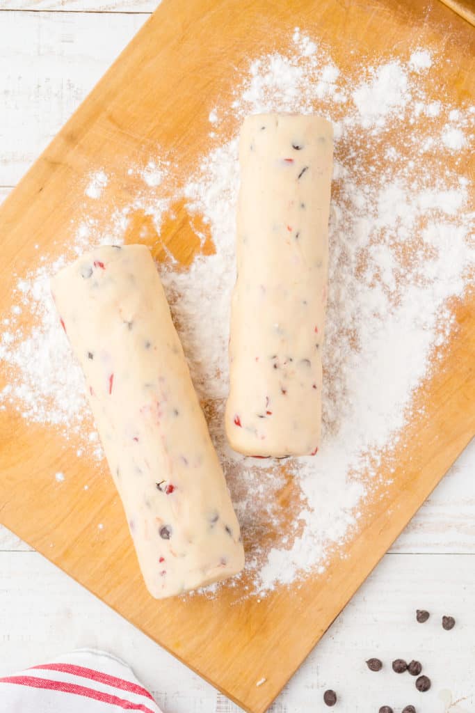 Two chocolate cherry shortbread dough rolled into logs on wood cutting board. 