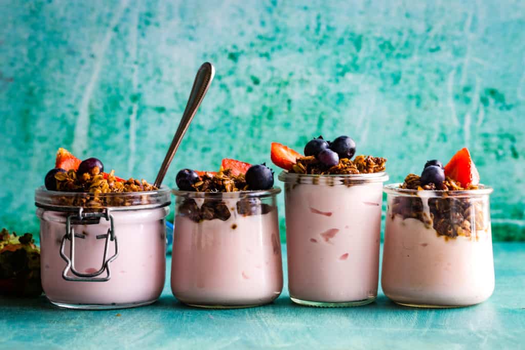 four jars of yogurt parfait with granola and berries lined up on a teal blue background. 