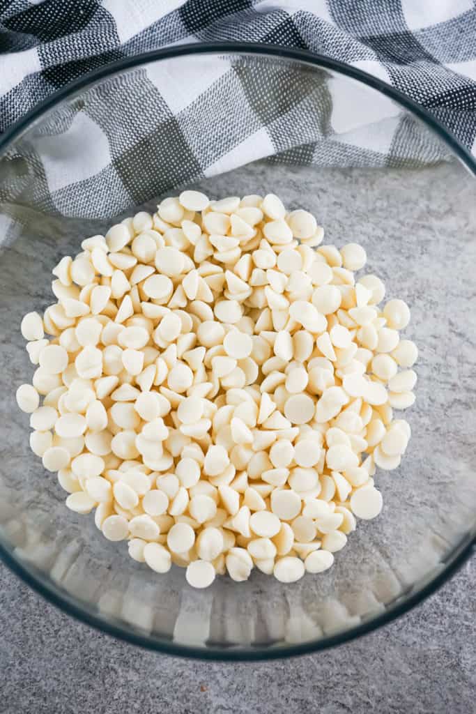 glass bowl full of white chocolate chips with black and white dish towel in background. 