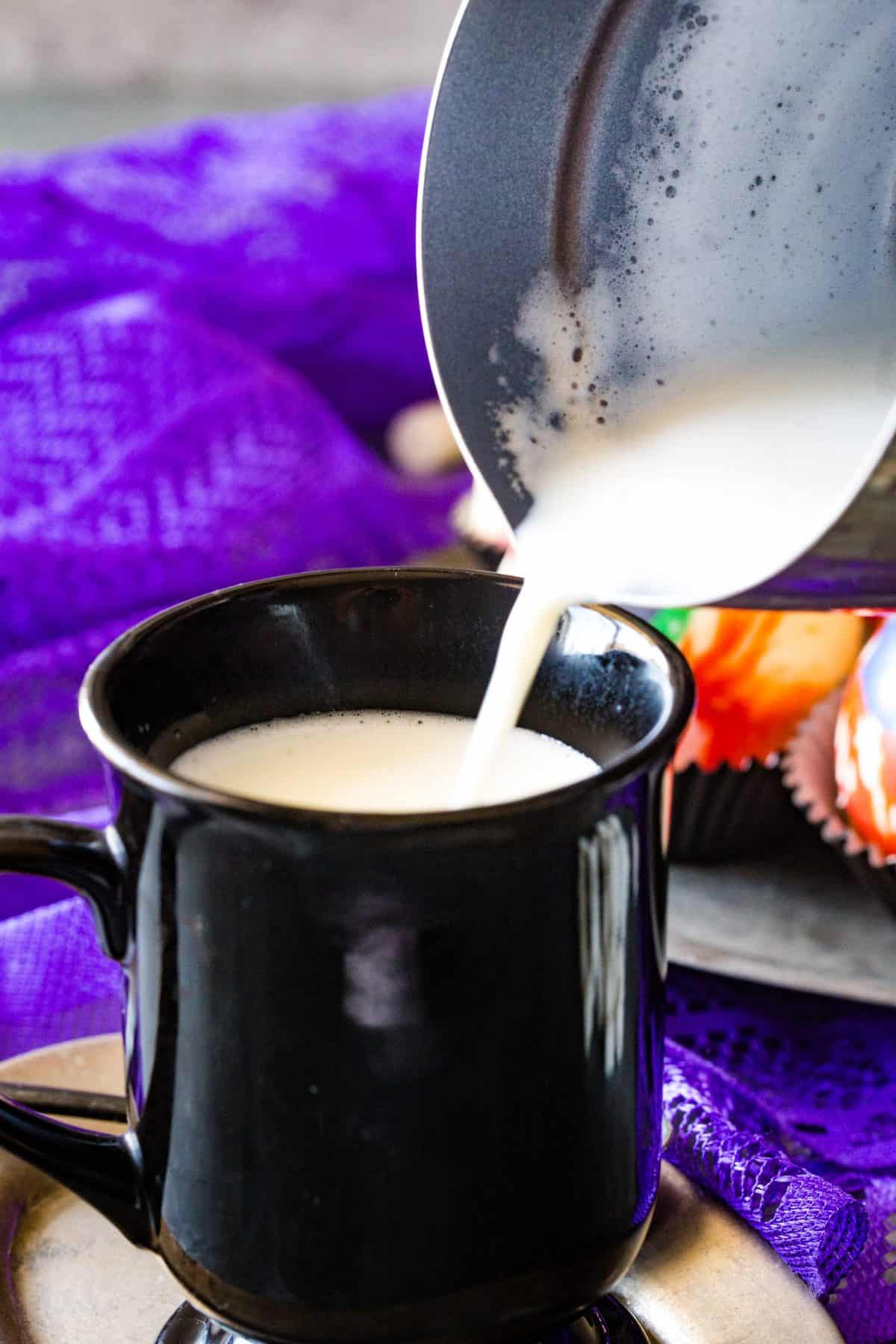 pouring the hot milk into the black coffee cup with purple towel in background. 