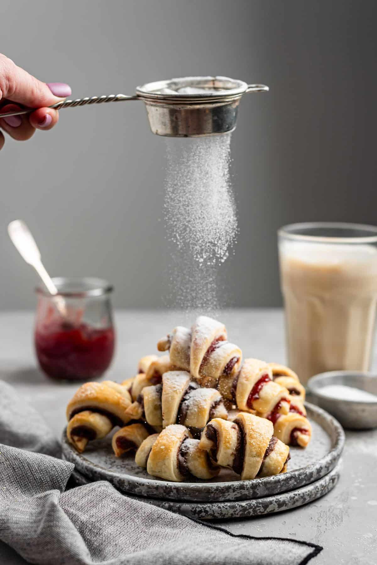 plate of rugelach cookies with a hand sift powdered sugar on top