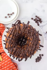 overhead shot of chocolate drizzled tiramisu bundt cake on white plate with orange dishtowel on the side