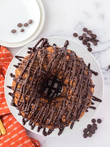 overhead shot of chocolate drizzled tiramisu bundt cake on white plate with orange dishtowel on the side