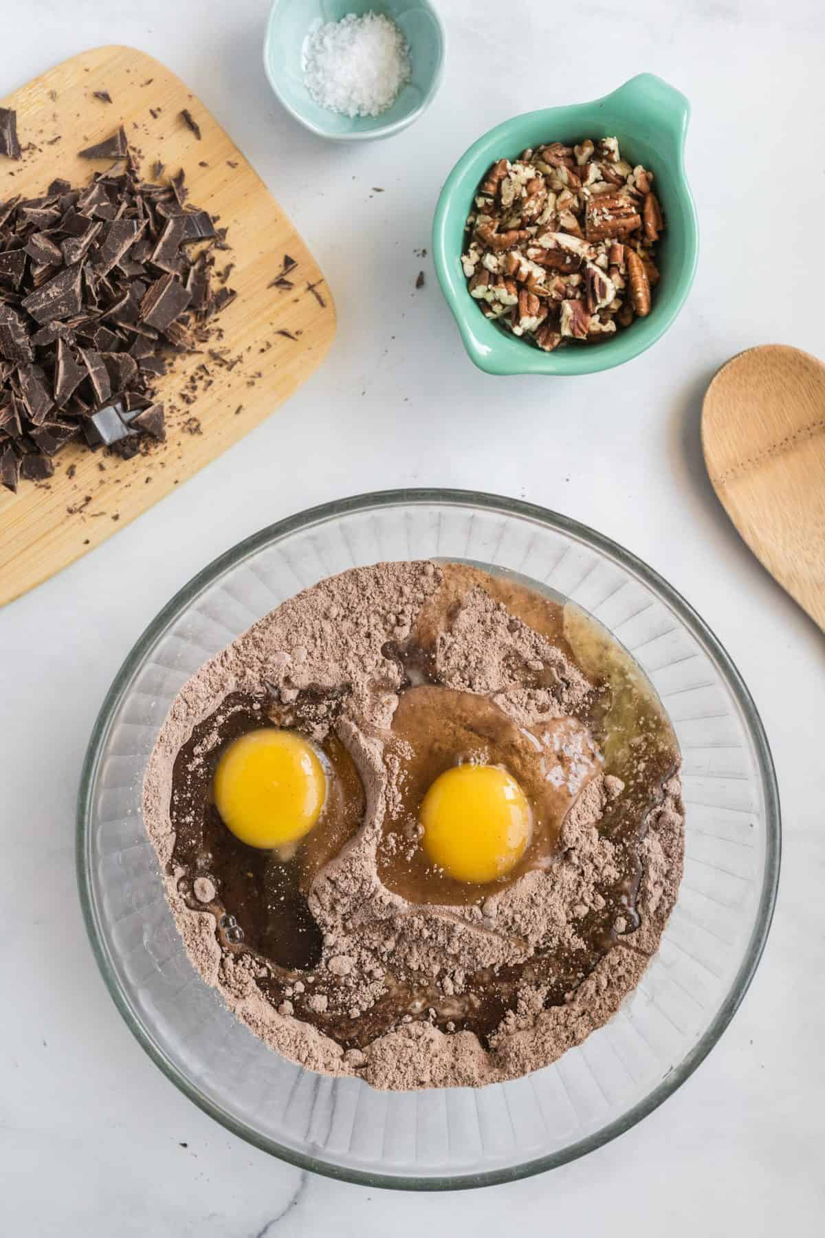 Cake mix and eggs in a large glass bowl with chocolate and pecans in smaller bowls.