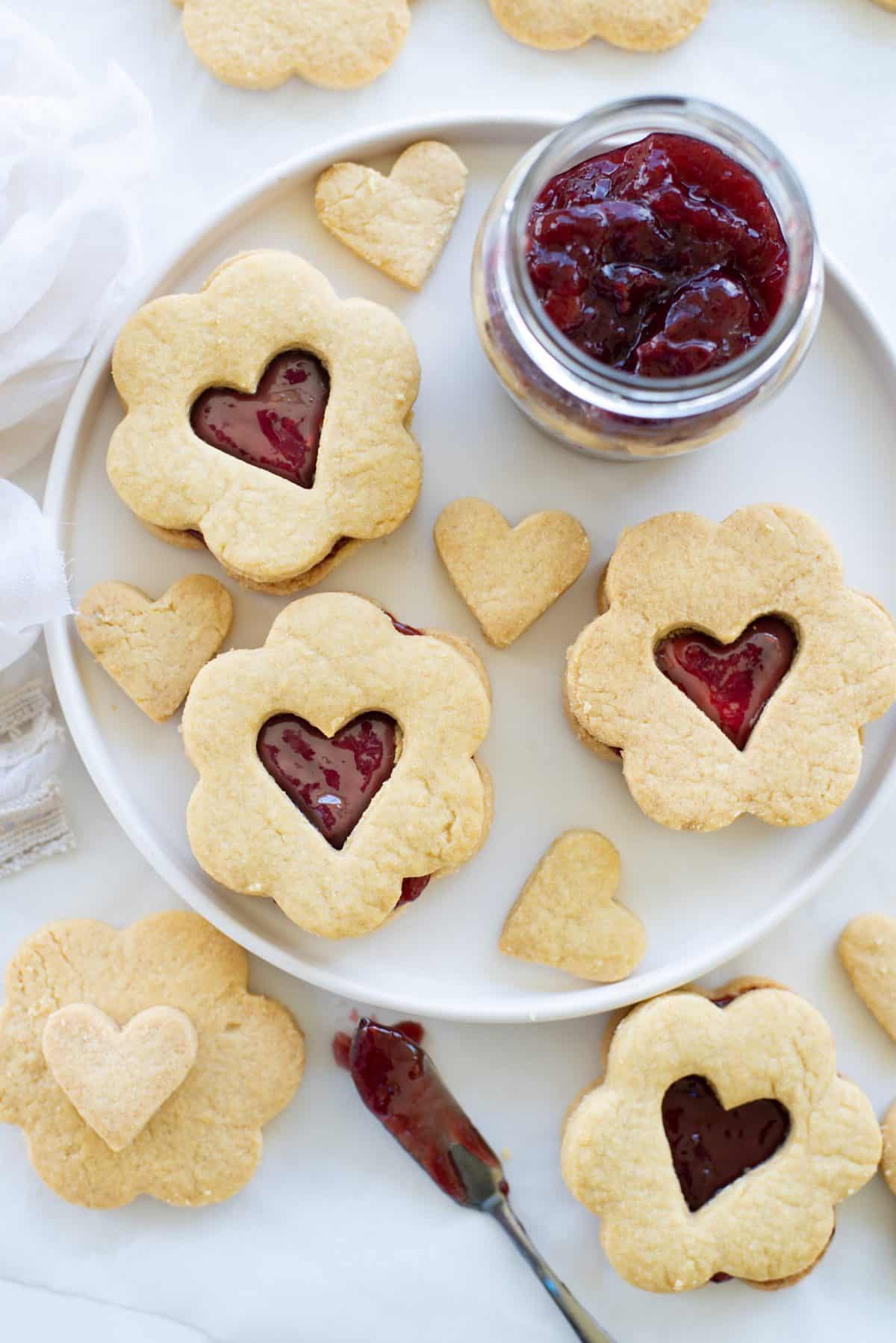 valentines day cookie sandwiches on white plate with a cup of jam next to them.
