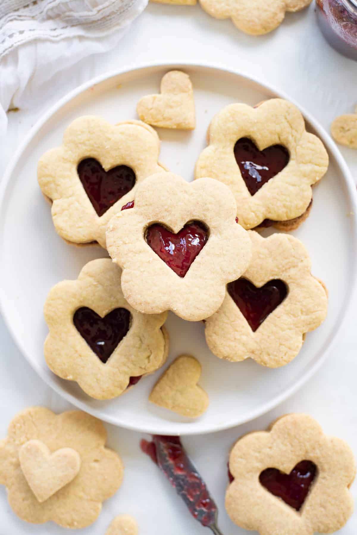 White plate with heart jam filled sugar cookie sandwiches on white background. 