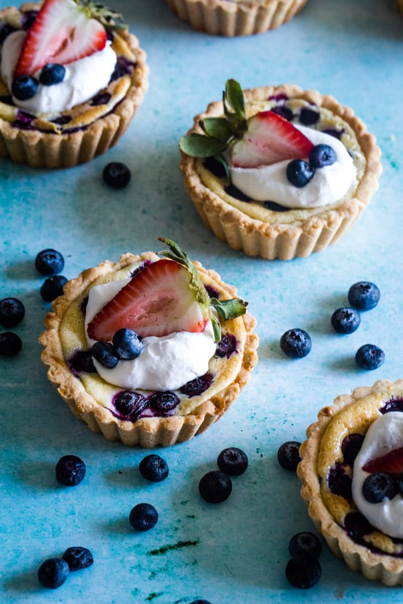 Mini cream cheese blueberry tarts on blue background with berries scattered. 