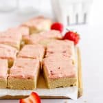 cutting board with sugar cookie bars and a white bowl with strawberries in background.