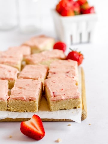 cutting board with sugar cookie bars and a white bowl with strawberries in background.
