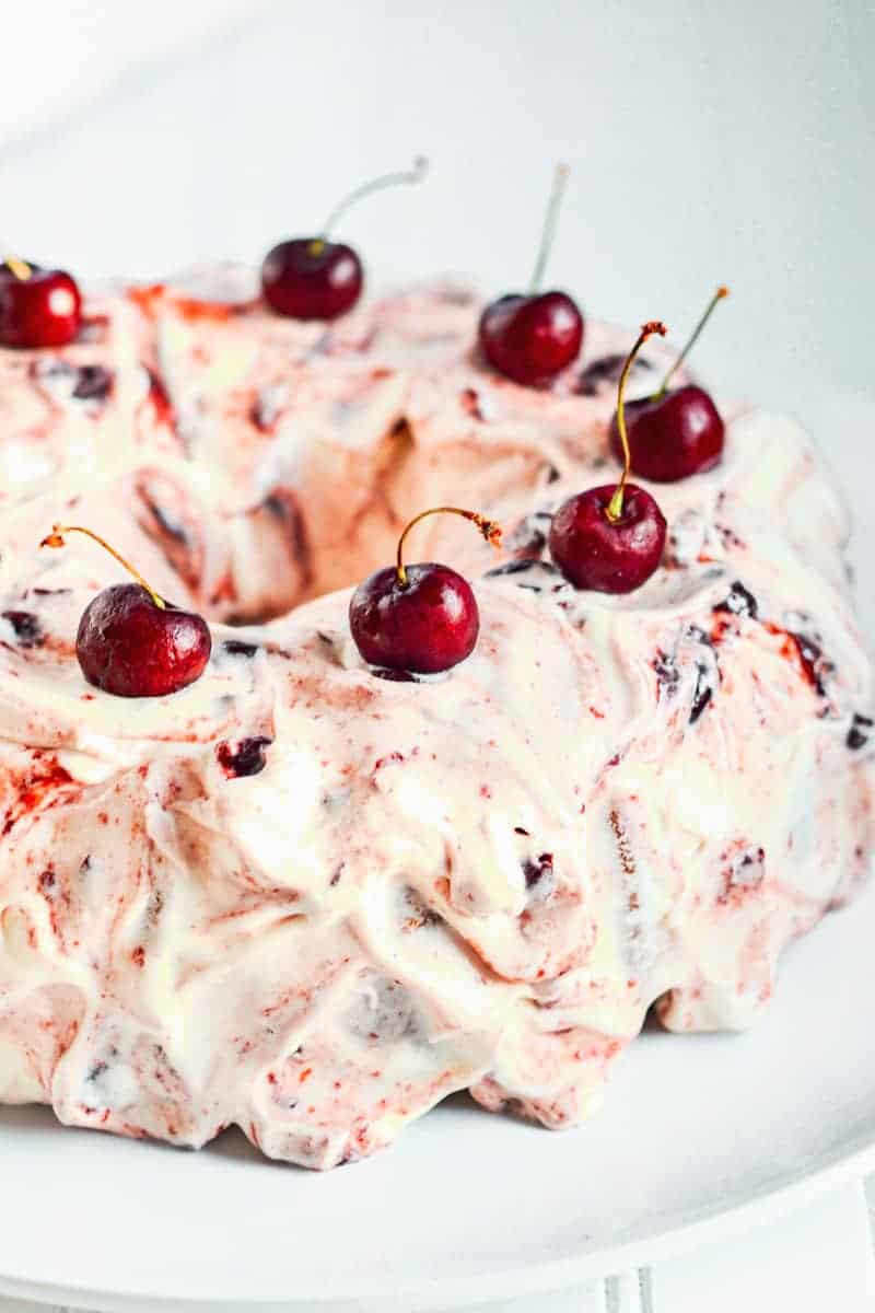 Bundt cake covered with cherry cream cheese frosting and fresh cherries on white plate.