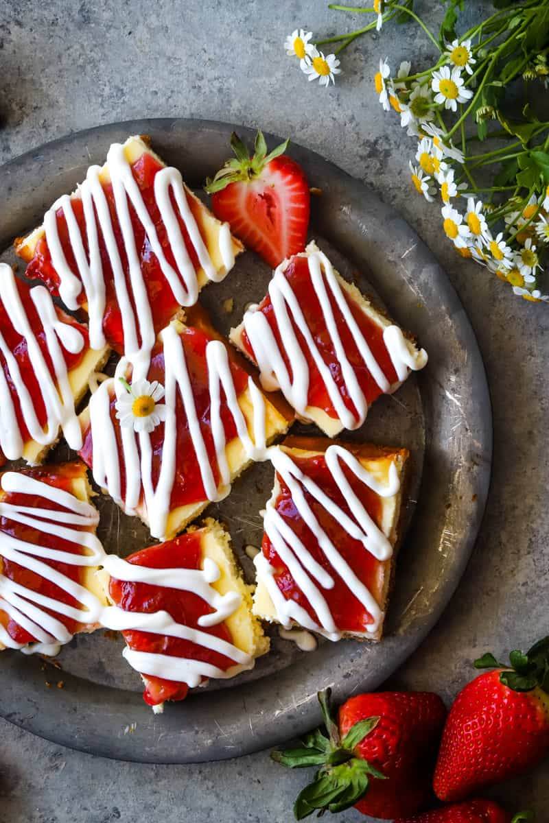 Plate of strawberry cheesecake bars with cut strawberries and flowers.