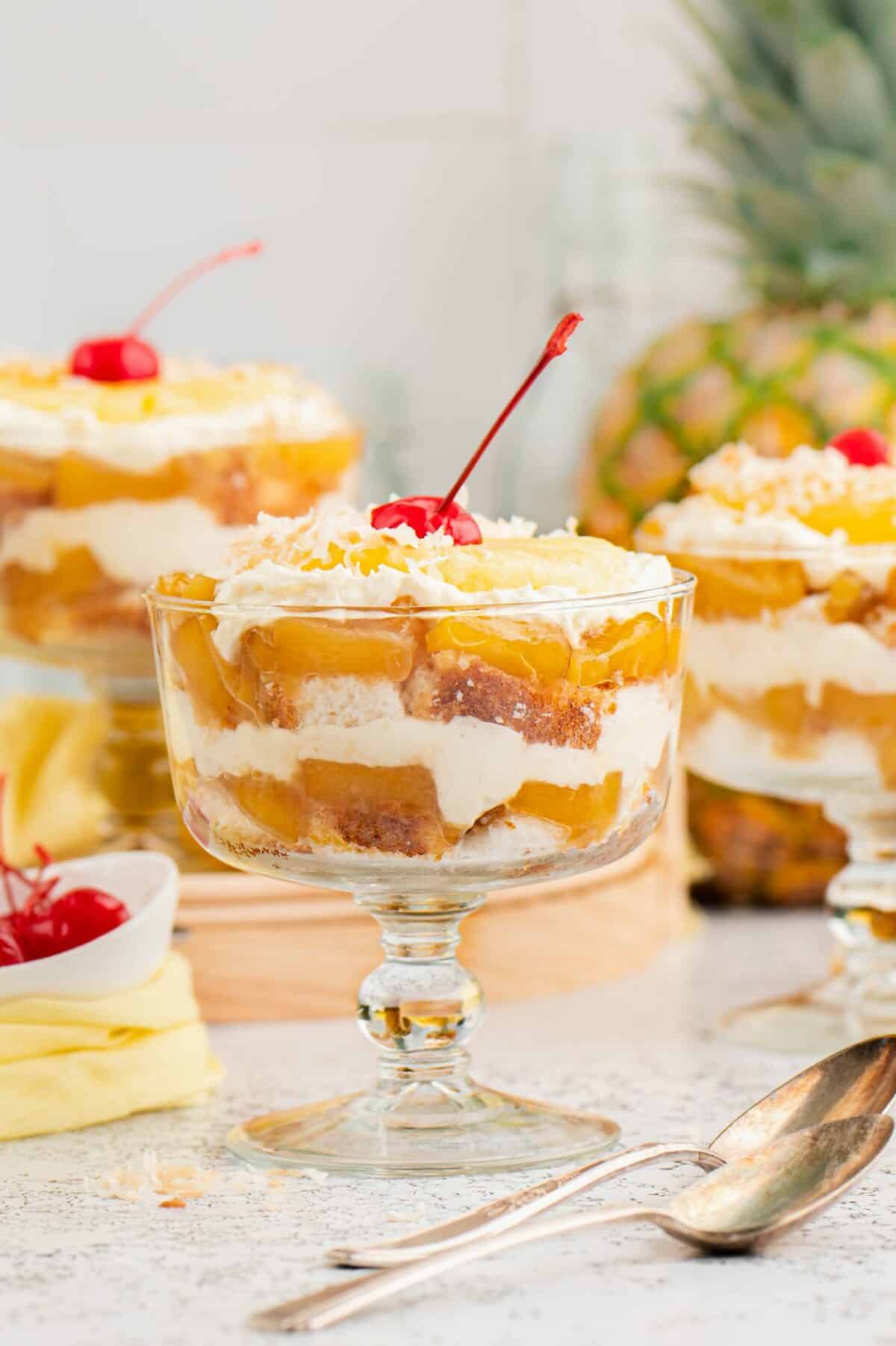 Three individual glasses with pineapple trifle and pineapple in the background. 