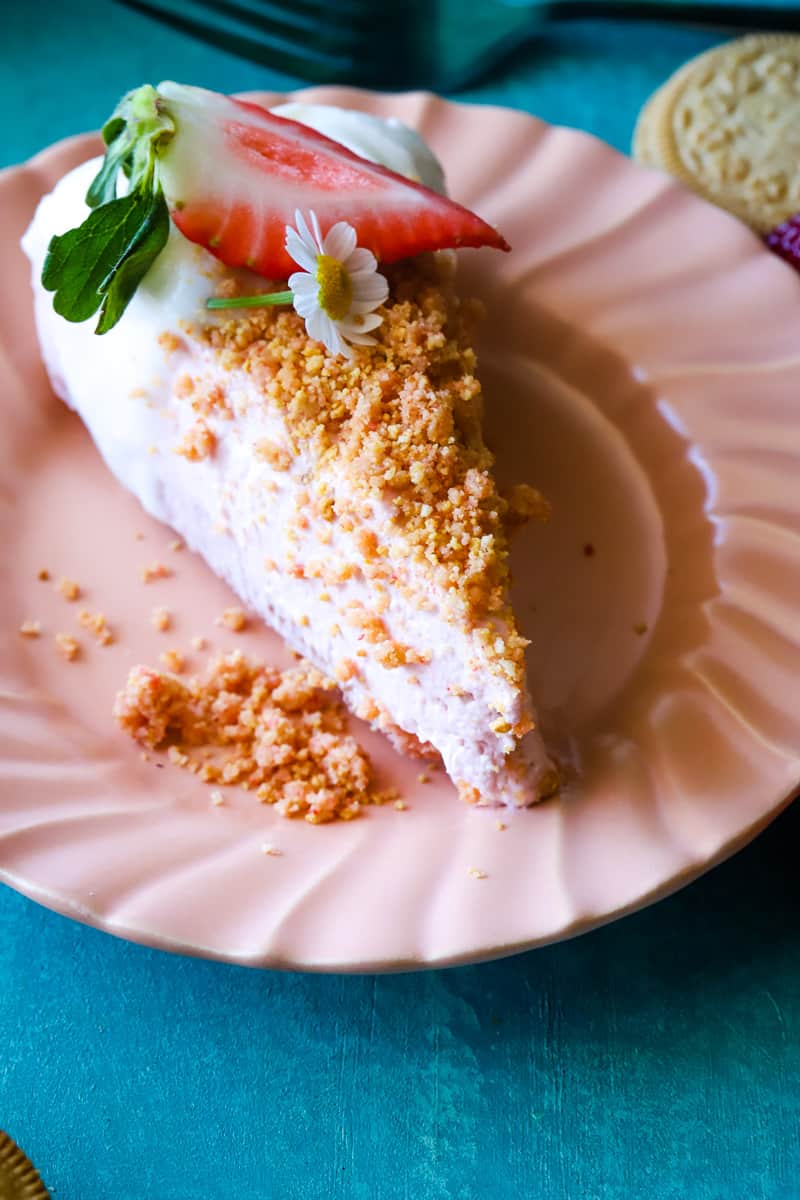 Slice of strawberry shortcake cheesecake on pink plate and blue backdrop.