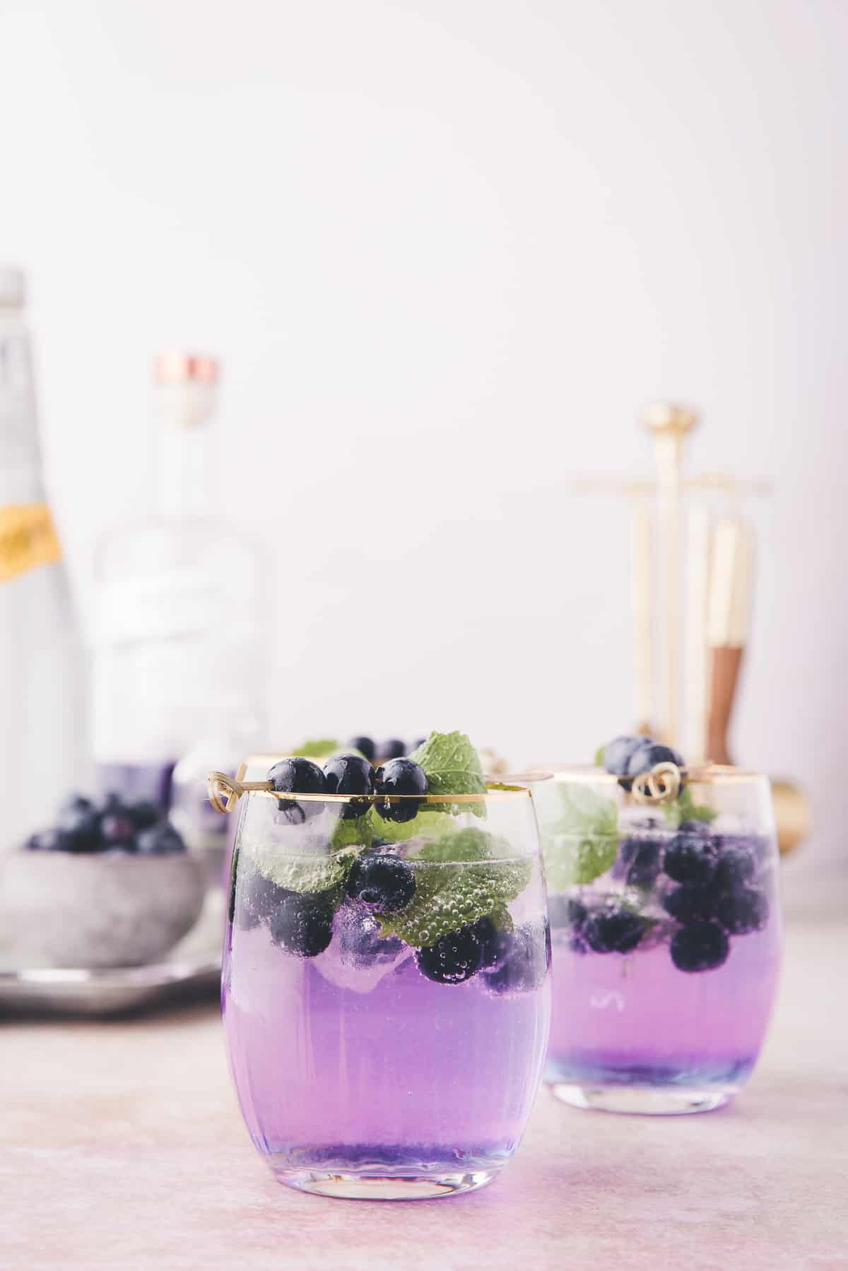 two purple cocktails with the ingredients to make the drinks in the background.