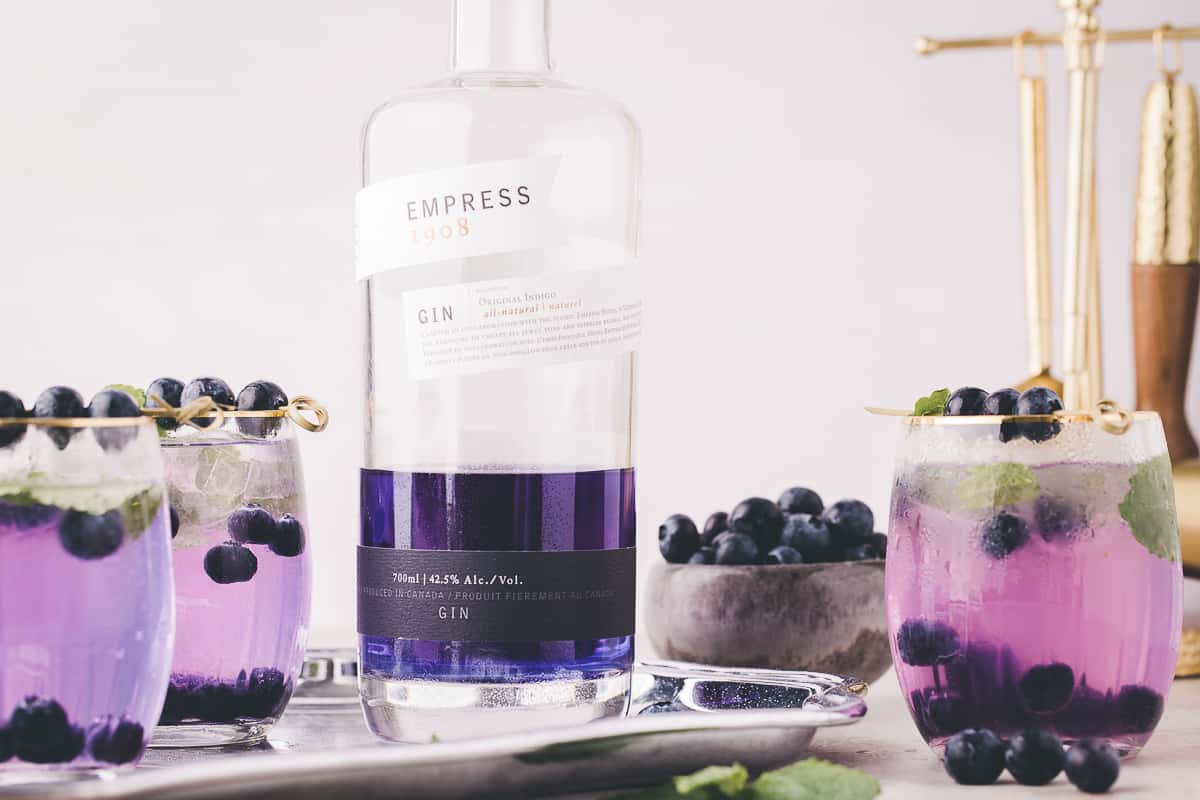 Bottle of empress gin with purple cocktails and fresh blueberries around it.