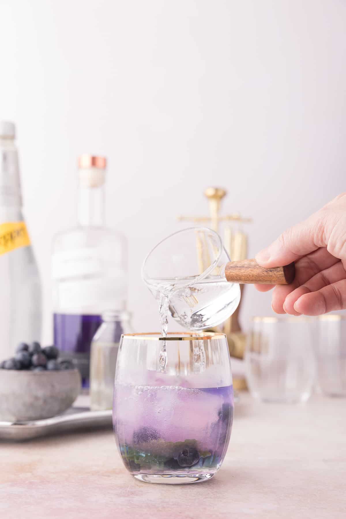 Hand pouring the soda water into the purple cocktail.