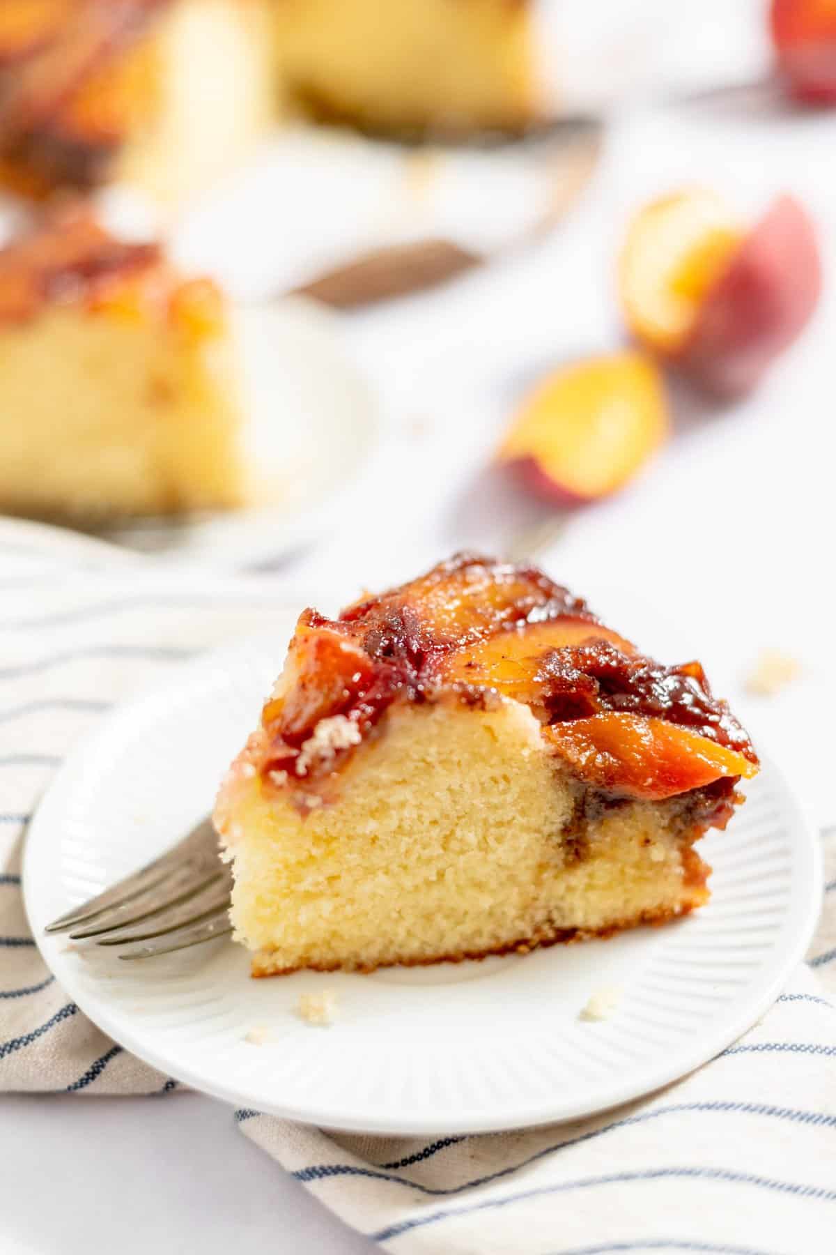 slice of cake on white plate with peaches blurred in background.