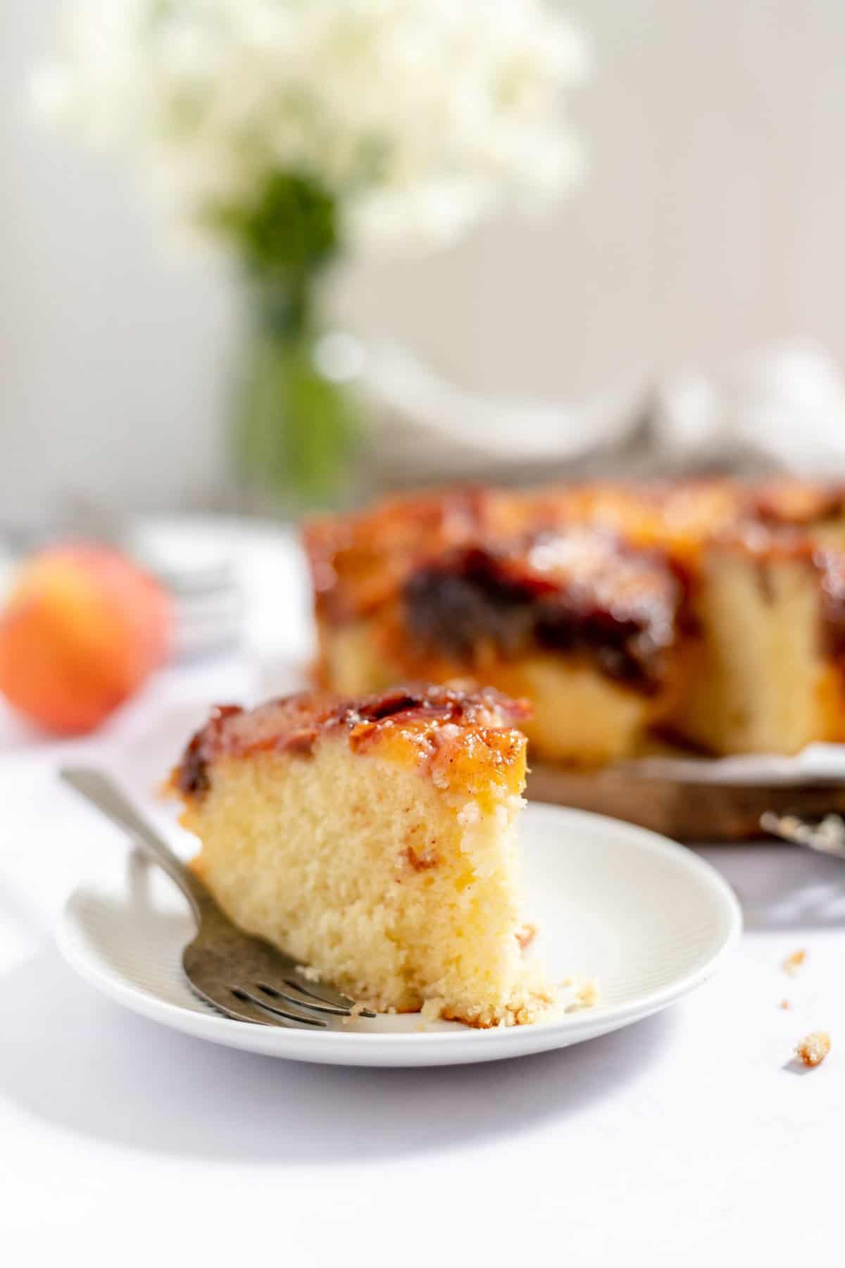 slice of cake on white plate with full cake and flowers in background. 